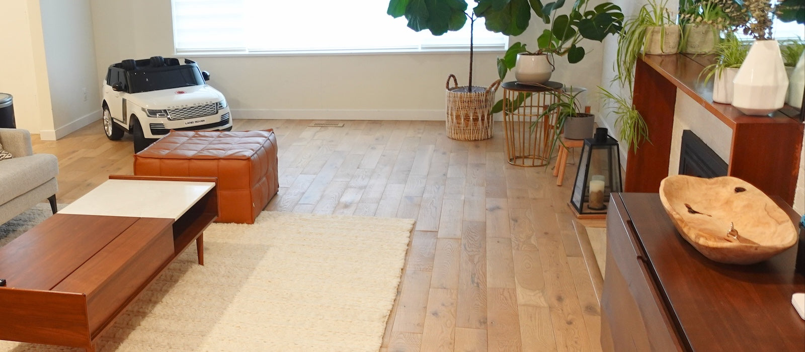 Cozy modern living room with natural oak hardwood flooring, warm-toned furniture, and indoor plants. The space features a fireplace, contemporary decor, and a neutral color palette for a relaxing ambiance.